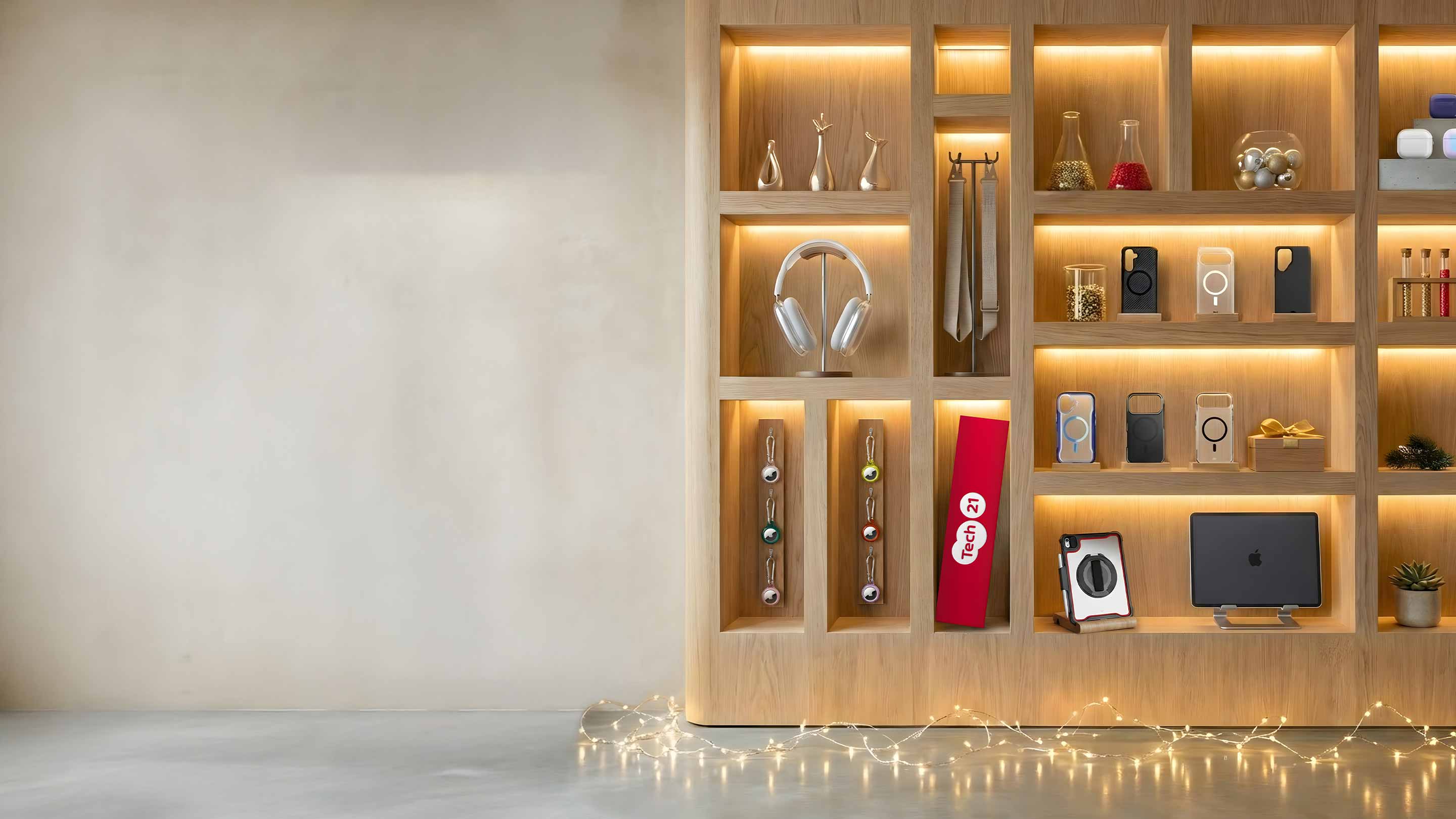 Various Tech21 products for iPhones, iPads, AirPods, and AirTag sit on lighted wood shelves with white Christmas lights on the floor