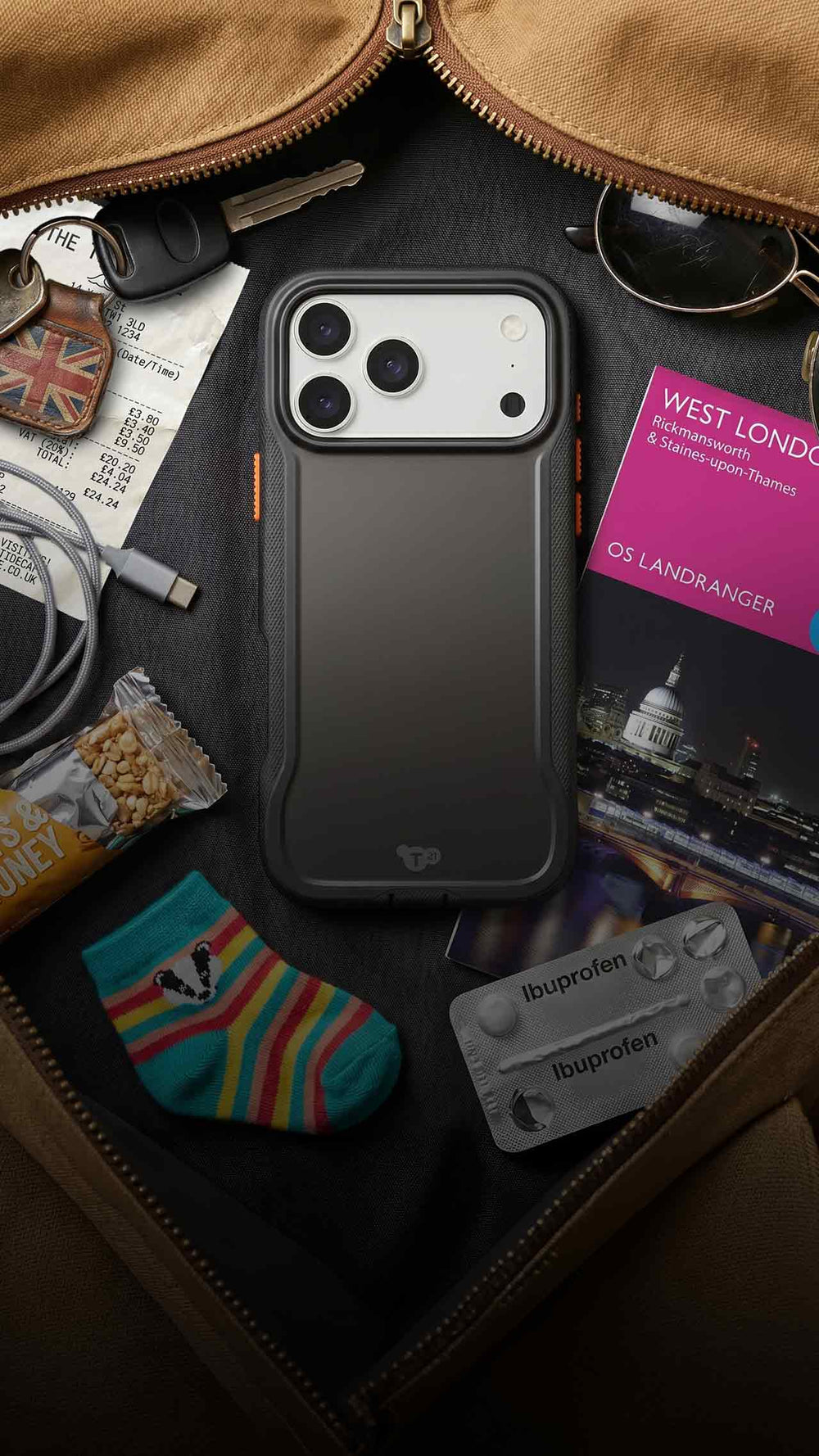 An iPhone in a case by Tech21 is shown in an open travel bag with keys, cables, snacks, and more around it