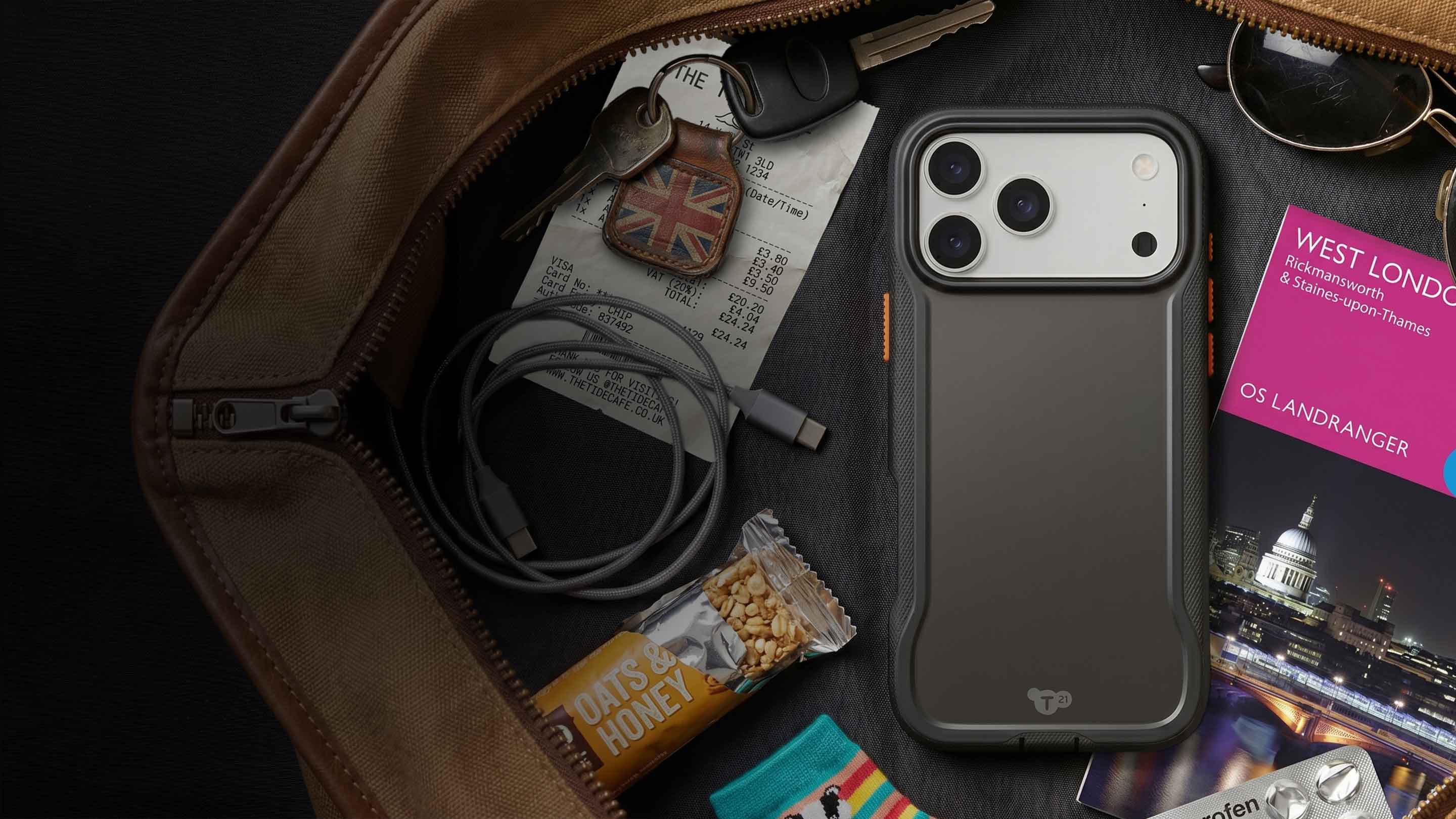 An iPhone in a case by Tech21 is shown in an open travel bag with keys, cables, snacks, and more around it